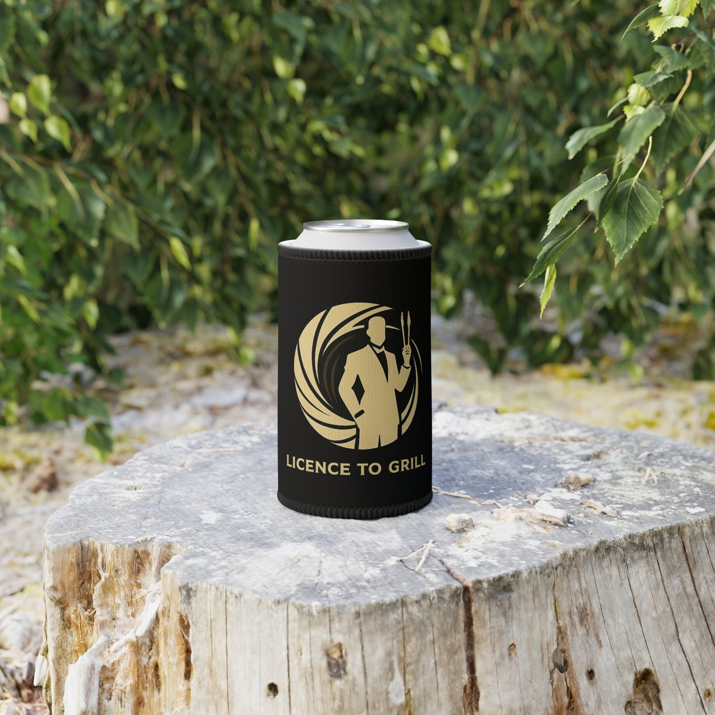 Black stubby holder with 'Licence to Grill' design on a wooden stump outdoors.